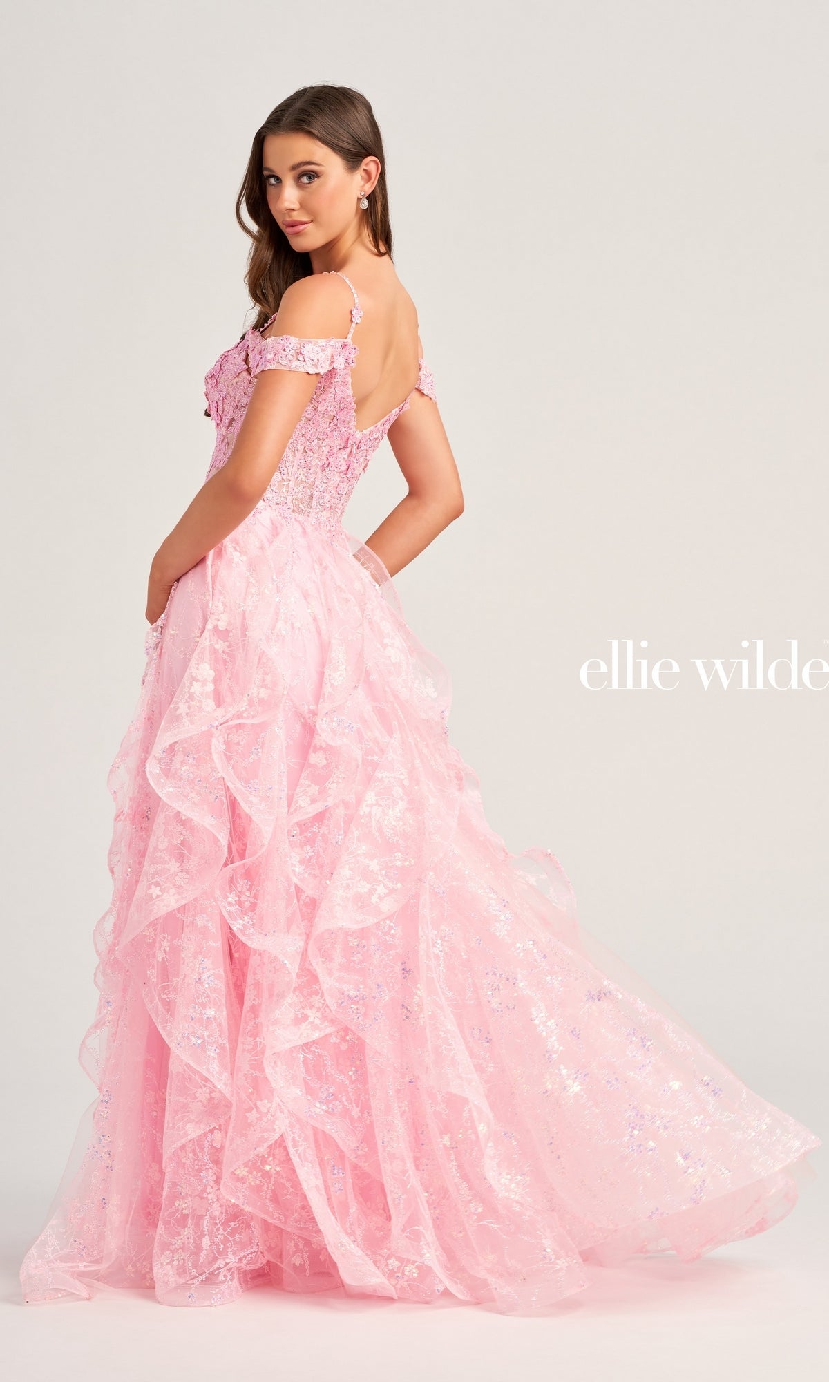 A woman poses with her back slightly turned, wearing the pink Ellie Wilde Ruffled Long Prom Ball Gown EW35218 against a plain background.